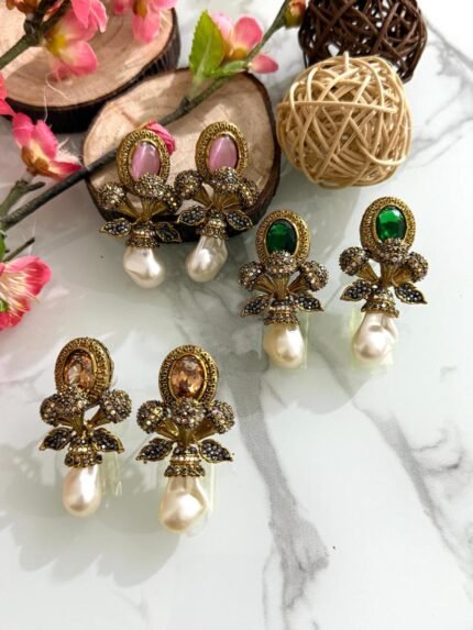 A set of floral-themed earrings with pearl drops and oval gemstones in pink, green, and amber, displayed on a white marble surface with decorative wooden and floral elements.