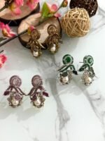 Three pairs of ornate earrings in gold, pink-silver, and green-silver tones with pearl drops and leaf motifs, displayed on a marble surface with floral and wooden decor.