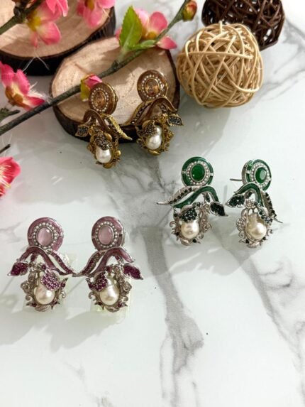 Three pairs of ornate earrings in gold, pink-silver, and green-silver tones with pearl drops and leaf motifs, displayed on a marble surface with floral and wooden decor.