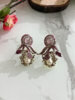Three pairs of ornate earrings in gold, pink-silver, and green-silver tones with pearl drops and leaf motifs, displayed on a marble surface with floral and wooden decor.