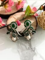 Three pairs of ornate earrings in gold, pink-silver, and green-silver tones with pearl drops and leaf motifs, displayed on a marble surface with floral and wooden decor.