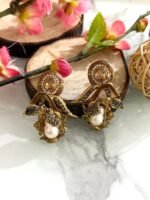 Three pairs of ornate earrings in gold, pink-silver, and green-silver tones with pearl drops and leaf motifs, displayed on a marble surface with floral and wooden decor.