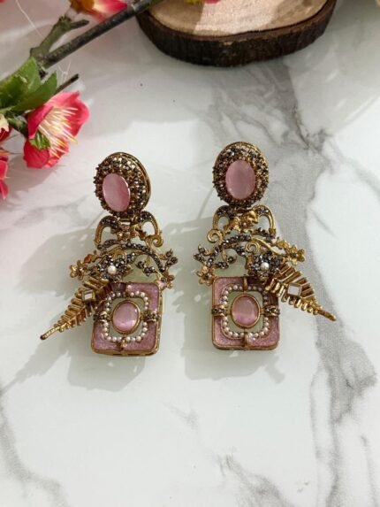 Pair of antique gold earrings with pink oval stones, floral detailing, pearls, and golden leaf motifs, displayed on a marble surface with floral accents.