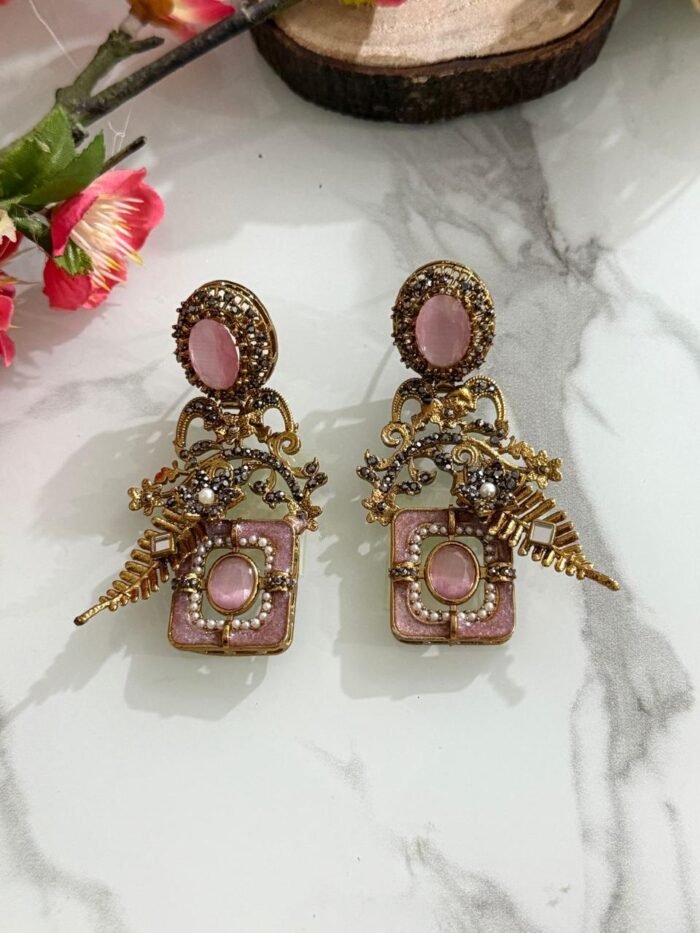 Pair of antique gold earrings with pink oval stones, floral detailing, pearls, and golden leaf motifs, displayed on a marble surface with floral accents.