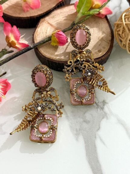 Pair of antique gold earrings with pink oval stones, floral detailing, pearls, and golden leaf motifs, displayed on a marble surface with floral accents.