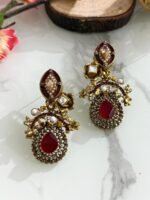 Pair of antique-style earrings with ruby red teardrop stones, kundan detailing, pearls, and gold floral designs on a marble background with wooden and floral props.