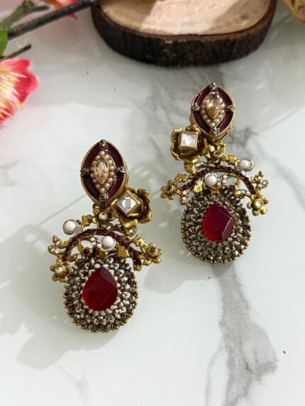 Pair of antique-style earrings with ruby red teardrop stones, kundan detailing, pearls, and gold floral designs on a marble background with wooden and floral props.