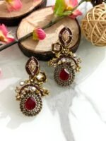 Pair of antique-style earrings with ruby red teardrop stones, kundan detailing, pearls, and gold floral designs on a marble background with wooden and floral props.