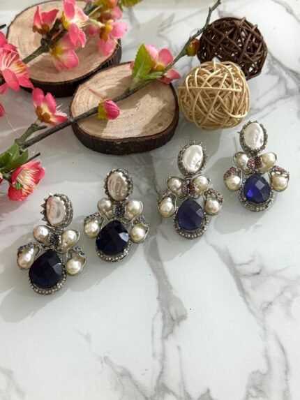 Three pairs of silver-finish earrings with blue teardrop stones and clustered pearls, placed on a marble surface with floral and wooden decor accents.
