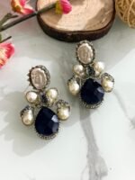 Three pairs of silver-finish earrings with blue teardrop stones and clustered pearls, placed on a marble surface with floral and wooden decor accents.