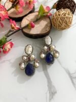 Three pairs of silver-finish earrings with blue teardrop stones and clustered pearls, placed on a marble surface with floral and wooden decor accents.