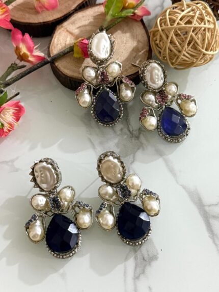 Three pairs of silver-finish earrings with blue teardrop stones and clustered pearls, placed on a marble surface with floral and wooden decor accents.