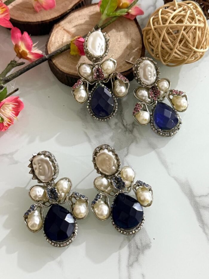 Three pairs of silver-finish earrings with blue teardrop stones and clustered pearls, placed on a marble surface with floral and wooden decor accents.