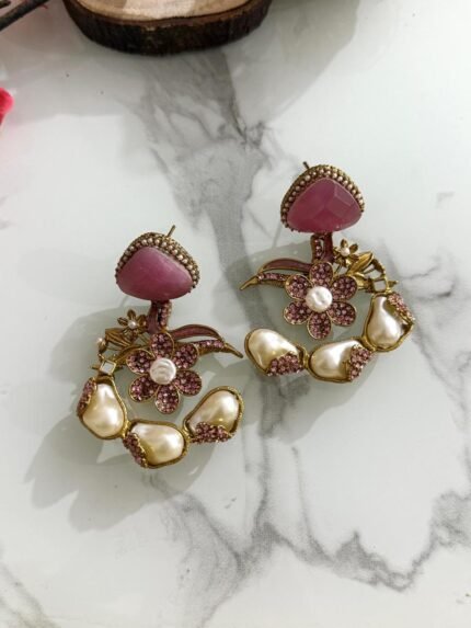 A pair of pink floral drop earrings with pearl accents and crystal detailing, displayed on a marble surface with a soft, elegant layout.