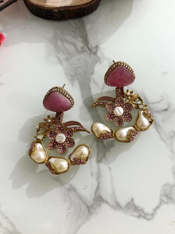 A pair of pink floral drop earrings with pearl accents and crystal detailing, displayed on a marble surface with a soft, elegant layout.