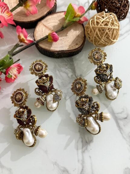 Antique-style drop earrings with rose motif and baroque pearls in maroon and black variants, displayed on a white marble surface with floral and wooden decor.