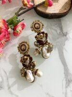 Antique-style drop earrings with rose motif and baroque pearls in maroon and black variants, displayed on a white marble surface with floral and wooden decor.
