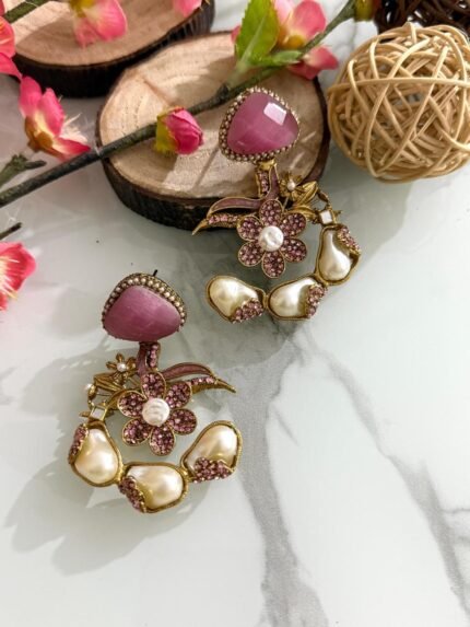 A pair of pink floral drop earrings with pearl accents and crystal detailing, displayed on a marble surface with a soft, elegant layout.