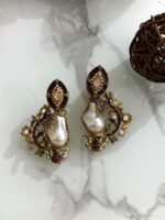 A pair of antique gold drop earrings with maroon enamel, crystals, and large baroque pearls placed on a marble surface with floral and wood props.