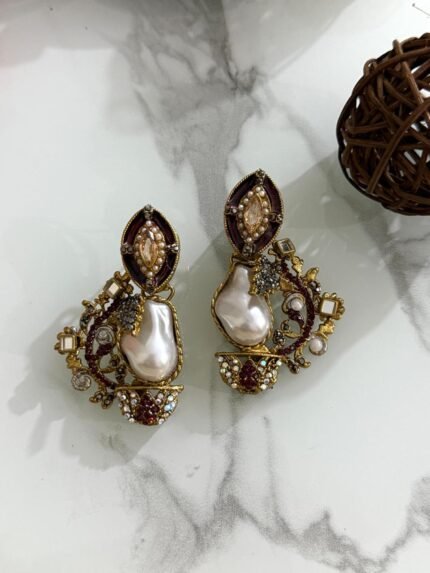A pair of antique gold drop earrings with maroon enamel, crystals, and large baroque pearls placed on a marble surface with floral and wood props.