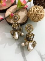 Antique-style drop earrings with rose motif and baroque pearls in maroon and black variants, displayed on a white marble surface with floral and wooden decor.