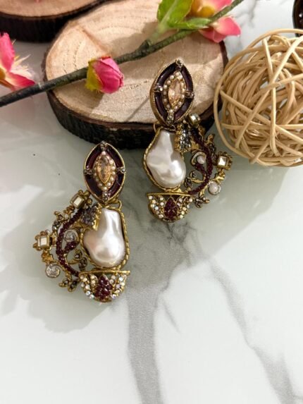 A pair of antique gold drop earrings with maroon enamel, crystals, and large baroque pearls placed on a marble surface with floral and wood props.