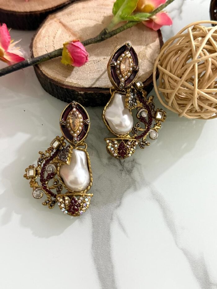 A pair of antique gold drop earrings with maroon enamel, crystals, and large baroque pearls placed on a marble surface with floral and wood props.