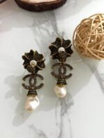 A pair of green crystal floral earrings with double C charm and hanging baroque pearl drops, displayed on a marble surface with floral and wooden decor.