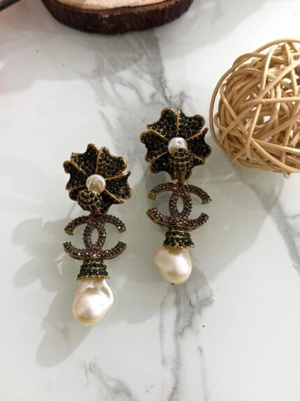 A pair of green crystal floral earrings with double C charm and hanging baroque pearl drops, displayed on a marble surface with floral and wooden decor.
