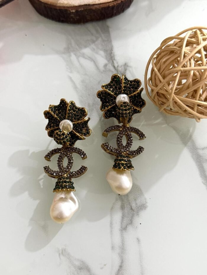 A pair of green crystal floral earrings with double C charm and hanging baroque pearl drops, displayed on a marble surface with floral and wooden decor.