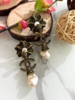 A pair of green crystal floral earrings with double C charm and hanging baroque pearl drops, displayed on a marble surface with floral and wooden decor.