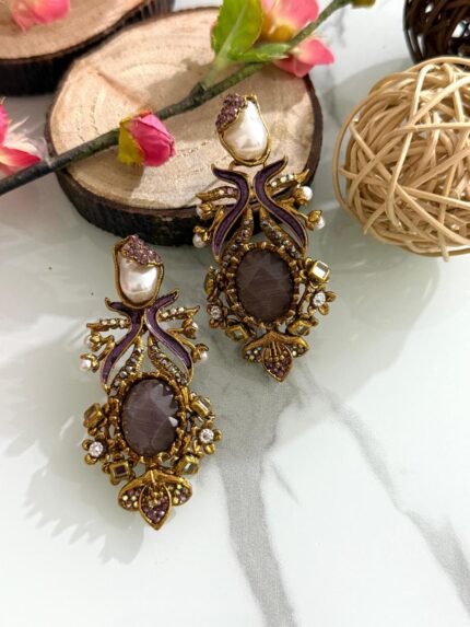 Pair of vintage-style earrings with purple enamel, central plum stone, pearl top, and antique gold detailing placed on a marble background with wooden and floral decor.