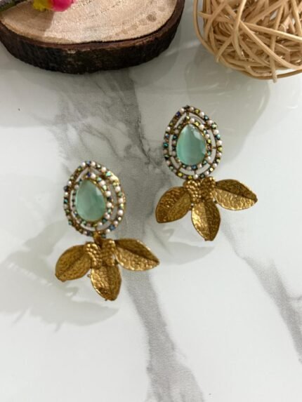 Earrings with mint green teardrop stones, pearl-studded border, and golden leaf design, displayed on a marble surface with floral and wooden decorations.