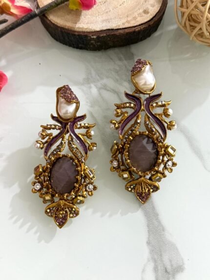 Pair of vintage-style earrings with purple enamel, central plum stone, pearl top, and antique gold detailing placed on a marble background with wooden and floral decor.