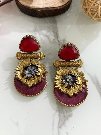 Earrings with red triangular stones, gold floral accents, and magenta velvet base featuring crystal embellishments, styled on a marble surface.