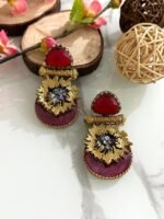 Earrings with red triangular stones, gold floral accents, and magenta velvet base featuring crystal embellishments, styled on a marble surface.