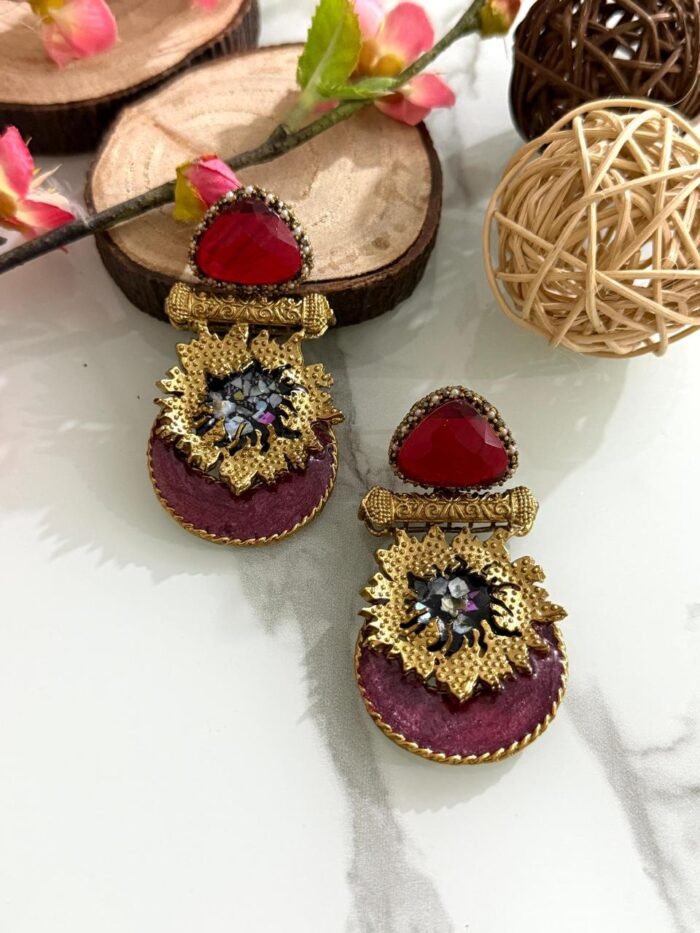 Earrings with red triangular stones, gold floral accents, and magenta velvet base featuring crystal embellishments, styled on a marble surface.