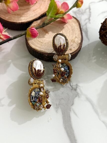 A pair of antique gold drop earrings with pearl tops, maroon flame detailing, and floral branches with pearls and stones, displayed on a decorative marble background.