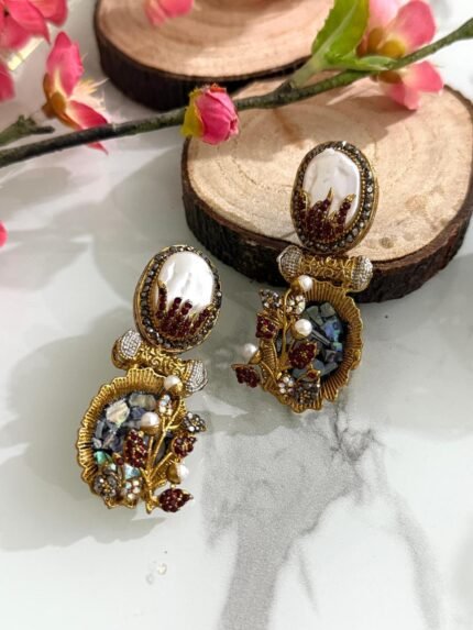 A pair of antique gold drop earrings with pearl tops, maroon flame detailing, and floral branches with pearls and stones, displayed on a decorative marble background.