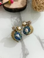 A pair of beaded stud earrings in red and blue with leaf motifs, pearl clusters, and central shell accents, displayed on a marble surface with floral props.