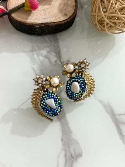 A pair of beaded stud earrings in red and blue with leaf motifs, pearl clusters, and central shell accents, displayed on a marble surface with floral props.