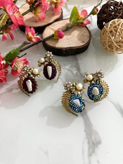A pair of beaded stud earrings in red and blue with leaf motifs, pearl clusters, and central shell accents, displayed on a marble surface with floral props.