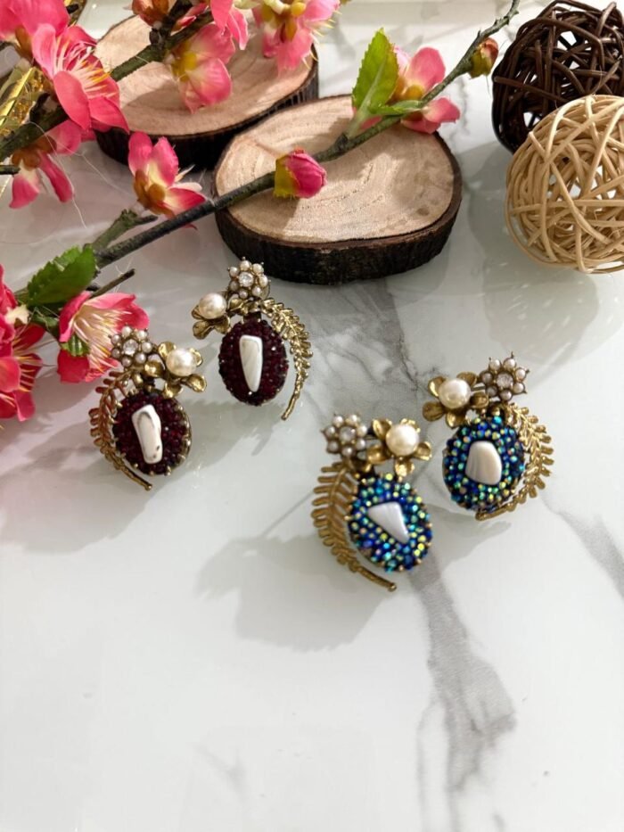 A pair of beaded stud earrings in red and blue with leaf motifs, pearl clusters, and central shell accents, displayed on a marble surface with floral props.
