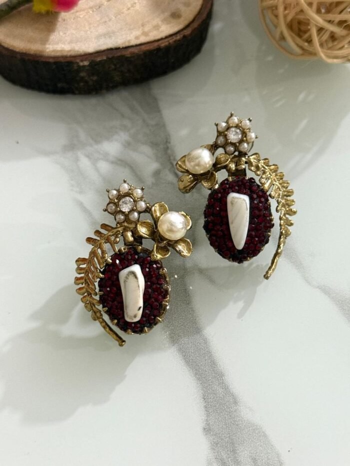 A pair of beaded stud earrings in red and blue with leaf motifs, pearl clusters, and central shell accents, displayed on a marble surface with floral props.