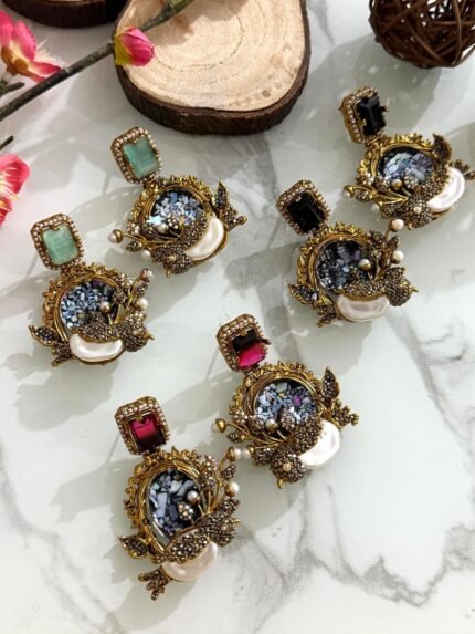 Six ornate earrings with square gemstone studs and circular mosaic centers, decorated with pearl clusters and antique gold detailing, displayed on a marble background.