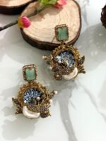 Six ornate earrings with square gemstone studs and circular mosaic centers, decorated with pearl clusters and antique gold detailing, displayed on a marble background.
