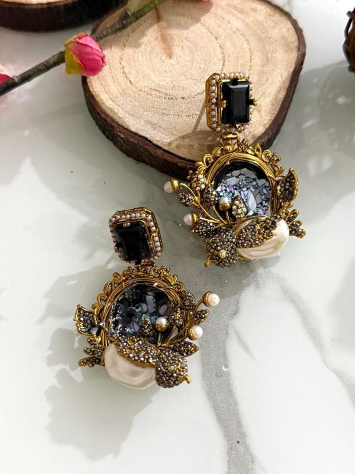 Six ornate earrings with square gemstone studs and circular mosaic centers, decorated with pearl clusters and antique gold detailing, displayed on a marble background.