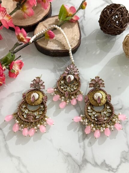 Antique gold maang tikka and earring set with kundan stones and pearl drop embellishments, displayed on a marble surface with floral and wooden decor accents.