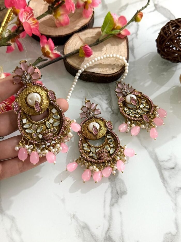 Antique gold maang tikka and earring set with kundan stones and pearl drop embellishments, displayed on a marble surface with floral and wooden decor accents.