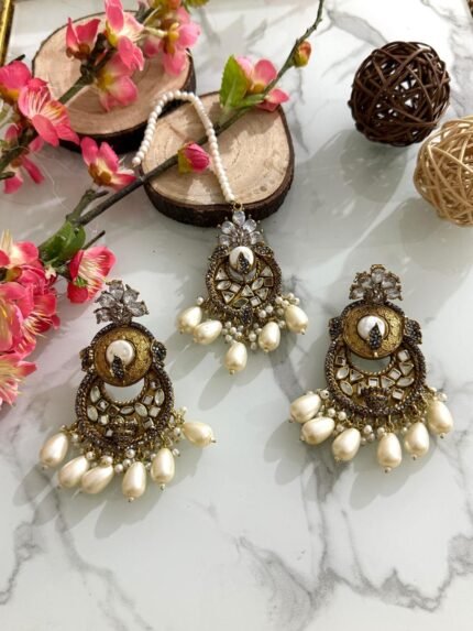 Antique gold maang tikka and earring set with kundan stones and pearl drop embellishments, displayed on a marble surface with floral and wooden decor accents.
